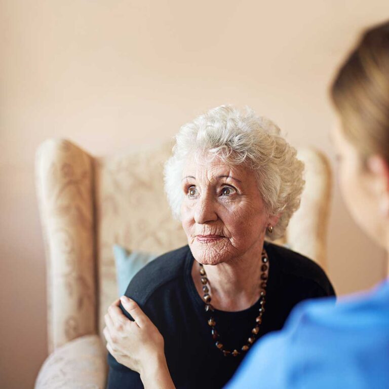 A senior woman sitting in a living room, looking thoughtful while a family member offers supportive conversation nearby.